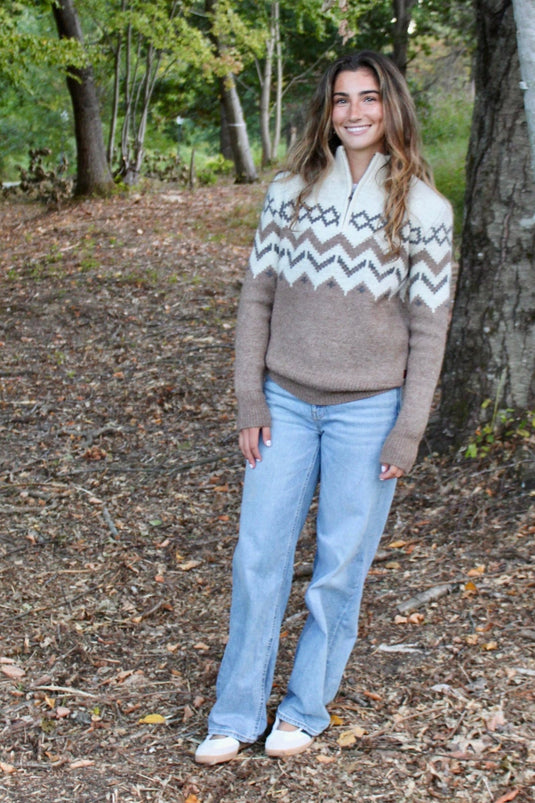 Woman standing in a forest wearing a patterned sweater and jeans.